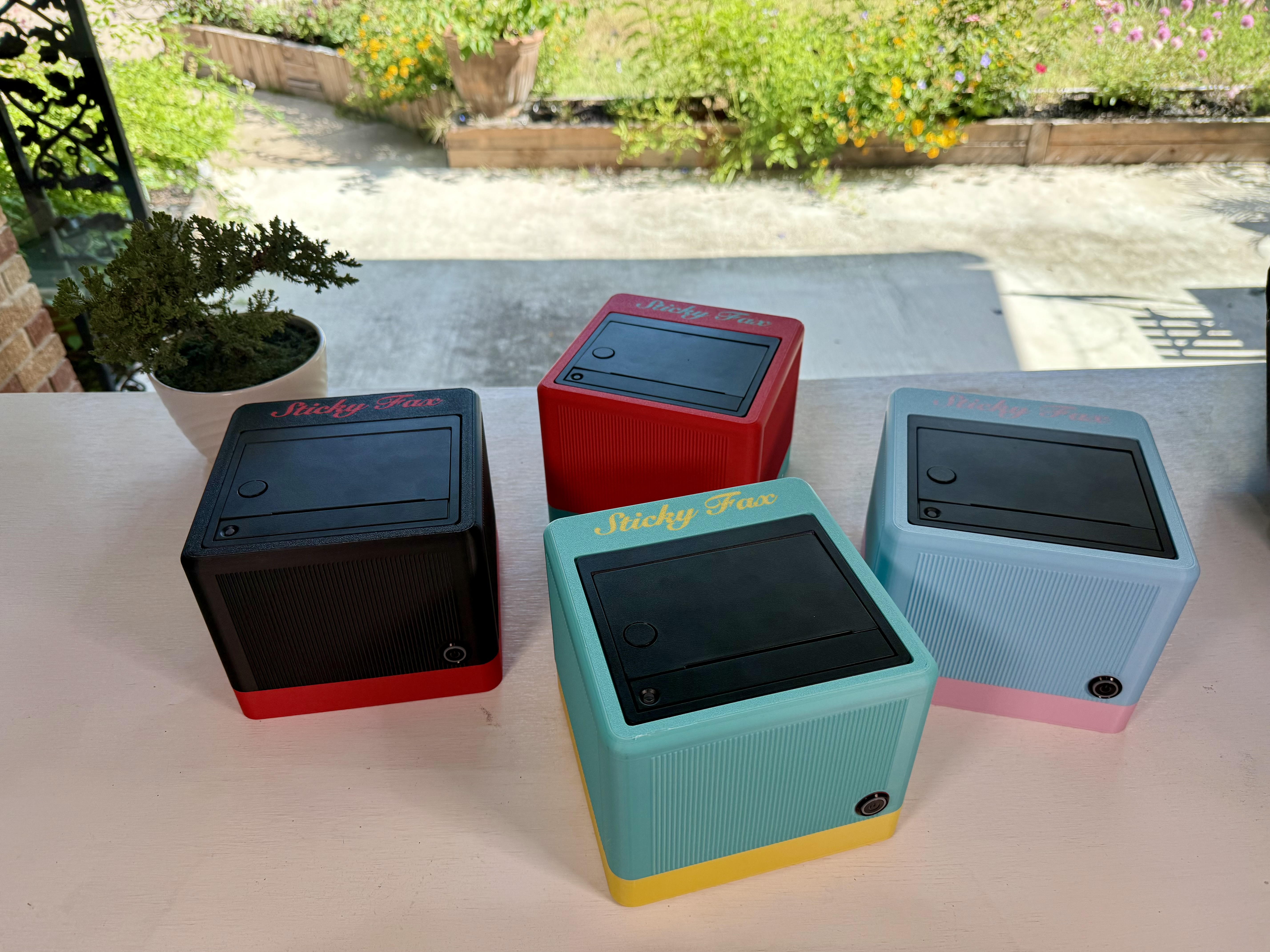 Four StickyFax units — black-and-red, red, teal-and-yellow, and blue-and-pink — on a table in a sunny garden
