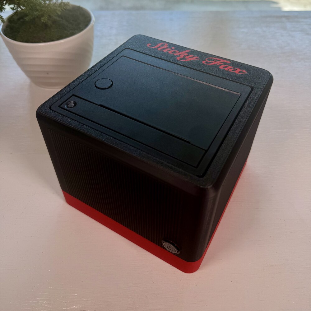 An ink-black-and-signal-red StickyFax on a desk, its power button lit