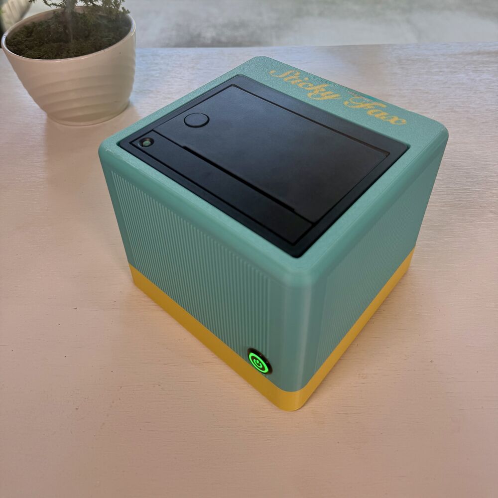 A teal-green-and-marigold StickyFax on a desk, power button glowing green